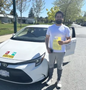 A bearded man in a white T-shirt and grey pants holds a yellow form and a black phone beside a white Seymour Driving School car with L and N student-driver magnets on the hood.