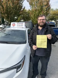 A bearded man in glasses, a black jacket, and dark jeans holds a yellow form beside a white Seymour Driving School car.
