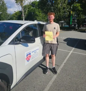 A graduate in a beige shirt holds a yellow form beside a white Seymour Driving School car.