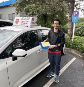 A graduate in a black jacket holds a yellow form beside a silver Seymour Driving School car.