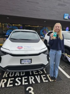 A graduate in a navy sweater holds a green N sign beside a white Seymour Driving School car.
