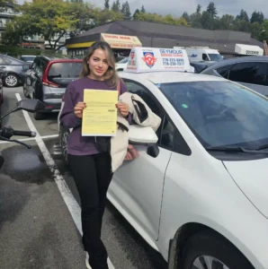 A graduate in a purple sweater holds a yellow form beside a white Seymour Driving School car.