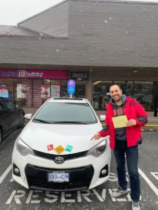 A graduate in a red jacket holds a yellow form beside a white car with learner and novice signs on the hood.