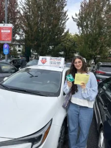 A graduate in a striped sweater holds a green N sign and a yellow form beside a white Seymour Driving School car.