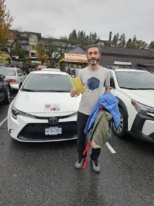 A man holds a yellow form, a blue jacket, and a backpack in front of a white Seymour Driving School car.