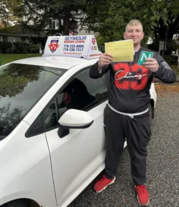 A man in a black Chicago jersey, grey pants, and red sneakers holds a yellow form and a green N sign beside a white Seymour Driving School car.