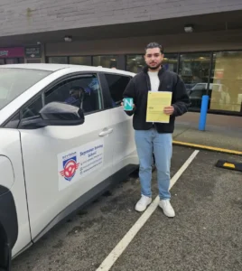 A man in a black fleece jacket, light blue jeans, and white sneakers holds a yellow form and a green N sign beside a white Seymour Driving School car outside a storefront.