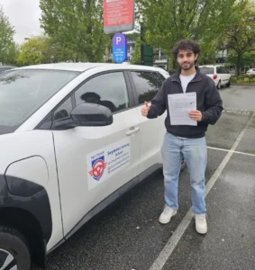 A man in a black hoodie and light blue jeans holds a printed document and gives a thumbs-up beside a white Seymour Driving School car.