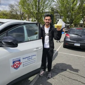 A man in a black jacket and white t-shirt holds a yellow form and papers beside the open door of a white Seymour Driving School car.