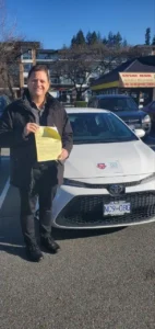 A man in a black jacket holds a yellow form in front of a white car in a sunny parking lot.