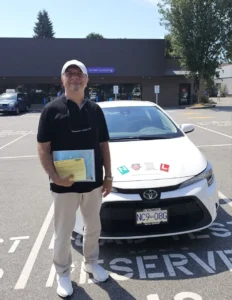 A man in a black polo and a white cap holds a yellow form in front of a white Seymour Driving School car.