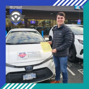A man in a black puffer jacket and blue jeans smiles while holding a yellow form beside a white Seymour Driving School car.