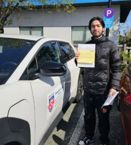 A man in a black puffer jacket holds a yellow form beside a white Seymour Driving School car near an ICBC road-test parking sign.