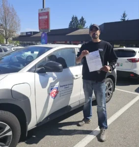 A man in a black sweatshirt, blue jeans, and a dark cap holds a white paper beside a white Seymour Driving School car in a sunny parking lot.