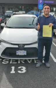 A man in a blue hoodie and a beanie gives a thumbs-up while holding a yellow form in front of a white car at a Driver Licensing lot.