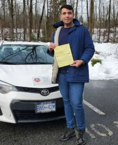 A man in a blue jacket and jeans holds a yellow form in front of a white Seymour Driving School car, with snow and bare trees behind him.