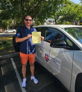 A man in a blue shirt, orange shorts, and white sneakers holds a yellow form beside a white Seymour Driving School car.
