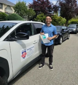 A man in a blue short-sleeve shirt and black pants holds papers and a green N sign beside a white Seymour Driving School car.
