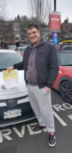 A man in a dark grey quilted jacket, purple hoodie, and grey sweatpants smiles while resting a yellow form on the hood of a white car in a parking lot.
