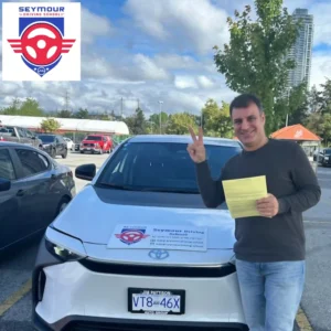 A man in a dark grey sweater and jeans holds a yellow form and makes a peace sign beside a white Seymour Driving School car.