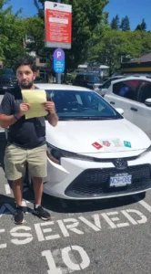 A man in a dark T-shirt and khaki shorts holds a yellow form in front of a white car with L and N signs on the hood.