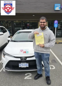 A man in a grey hoodie, glasses, and blue jeans holds a yellow form in front of a white Seymour Driving School car.