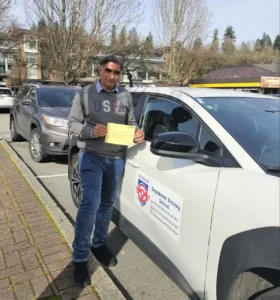 A man in a grey sweater and blue jeans holds a yellow form with both hands beside a white Seymour Driving School car parked along a brick sidewalk.