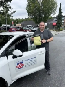 A man in a grey t-shirt and black pants holds a yellow form beside the open door of a white Seymour Driving School car.
