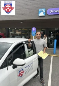 A man in a grey T-shirt holds a yellow form and gives a thumbs-up beside a white Seymour Driving School car outside an ICBC Driver Licensing office.