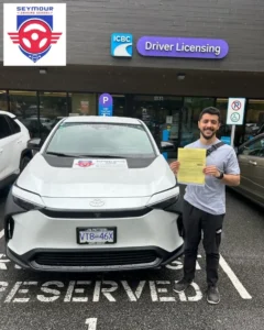 A man in a grey T-shirt holds a yellow form beside a white Seymour Driving School car outside an ICBC Driver Licensing office.