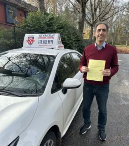 A man in a maroon sweater and dark jeans holds a yellow form beside a white Seymour Driving School car.
