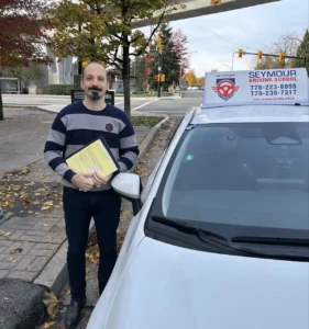 A man in a navy-and-grey striped sweater and black pants holds a yellow form on a clipboard beside a white Seymour Driving School car.