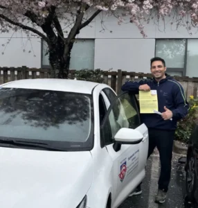 A man in a navy blue track jacket and black pants smiles while leaning on the open door of a white Seymour Driving School car and holding a yellow form and a pen under pink cherry blossoms.