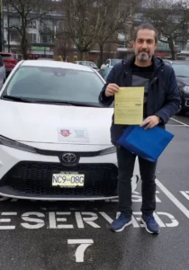 A man in a navy jacket holds a yellow form and a blue folder in front of a white Seymour Driving School car.
