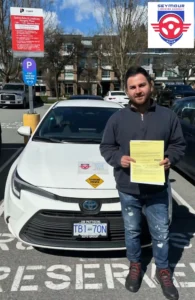 A man in a navy pullover and blue jeans holds a yellow form beside a white Seymour Driving School car in a parking lot.
