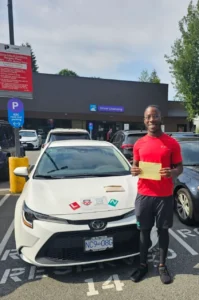 A man in a red t-shirt and black shorts smiles while holding a yellow form beside a white car with green N and red L signs on the hood outside a driver licensing office.