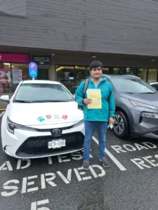 A man in a teal jacket and blue jeans holds a yellow form in front of a white Seymour Driving School car with green N, crest, and red L stickers on the hood.