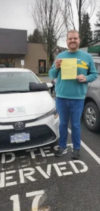 A man in a teal sweatshirt and blue jeans smiles while holding a yellow form in front of a white car in a reserved parking stall.