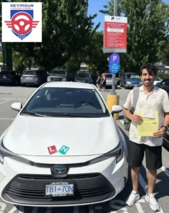 A man in a white patterned shirt and black shorts holds a yellow form beside a white Seymour Driving School car in a sunny parking lot.