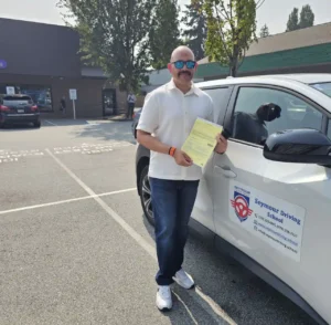 A man in a white polo, blue jeans, and sunglasses holds a yellow form beside a white Seymour Driving School car.
