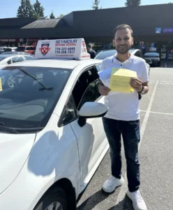 A man in a white polo, dark jeans, and white sneakers holds a stack of yellow forms beside a white Seymour Driving School car.