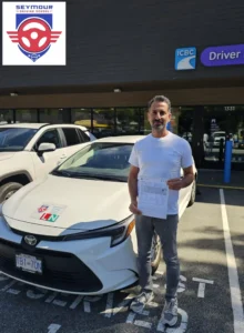 A man in a white T-shirt and grey pants holds a white form in front of a white Seymour Driving School car outside an ICBC driver licensing office.