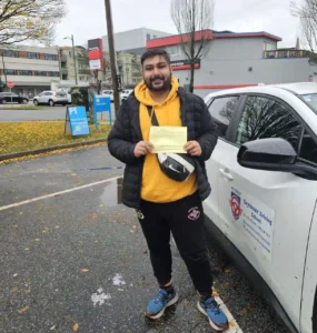A man in a yellow hoodie, a black puffer jacket, and blue sneakers holds a yellow form beside a white Seymour Driving School car.