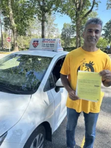 A man in a yellow T-shirt and blue jeans holds a yellow form beside a white Seymour Driving School car.