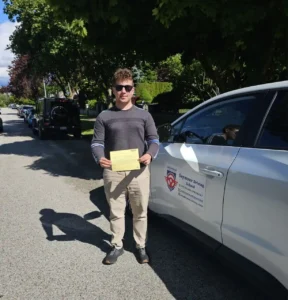 A man in sunglasses, a grey striped sweater, and beige pants holds a yellow form beside a white Seymour Driving School car on a tree-lined street.
