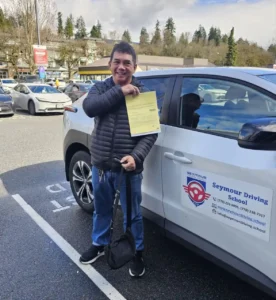 A smiling man in a black puffer jacket and blue jeans holds a yellow form beside a white Seymour Driving School car.