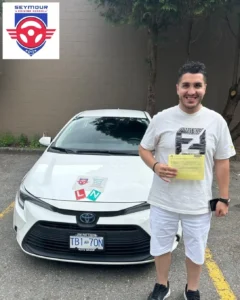 A smiling man in a white graphic T-shirt and white shorts holds a yellow form in front of a white car with red L and green N signs on the hood.