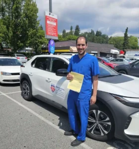 A smiling man in blue scrubs holds a yellow form beside a white Seymour Driving School car in a sunny parking lot.