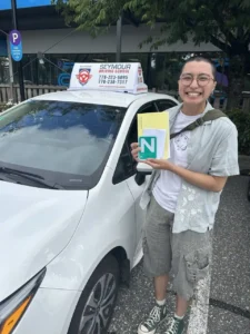 A smiling person with short dark hair and glasses, wearing a pale grey shirt and grey cargo shorts, holds a yellow form and a green N sign beside a white Seymour Driving School car with a roof sign.