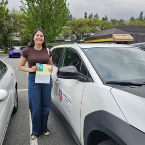 A smiling woman in a brown T-shirt and blue jeans holds a yellow form and a green N sign next to a white Seymour Driving School car.